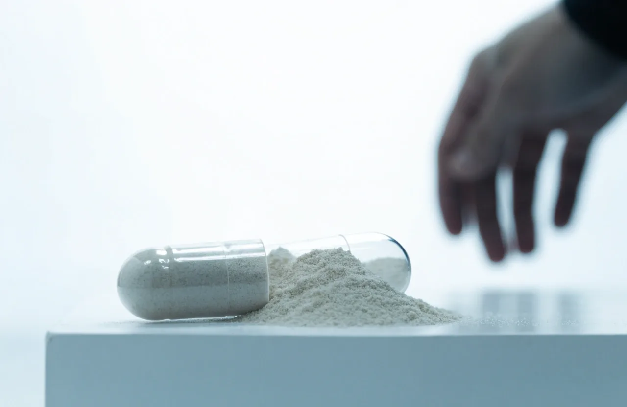 A split clear capsule with pale powder spilled on a white plinth, an out-of-focus hand at the frame edge